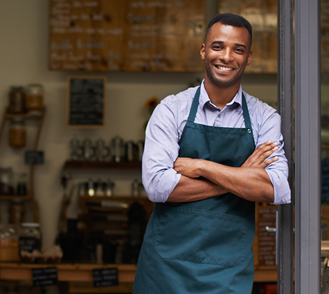 Image of young Black business owner wearing an apron and standing in shop doorway.