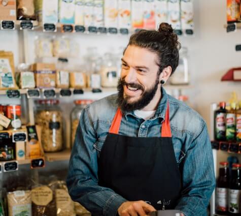 Young hipster business owner with bun and earring wearing an apron and smiling as he swipes a credit card.