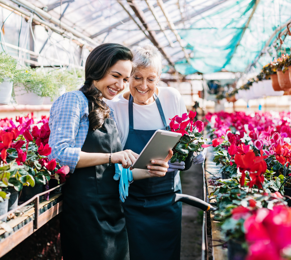 Two women looking at a tablet while one holds flowers in the greenhouse of their florist shop.