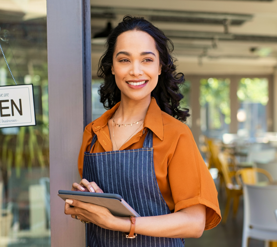 Female business owner stands outside her restaurant wearing an apron and holding a tablet.