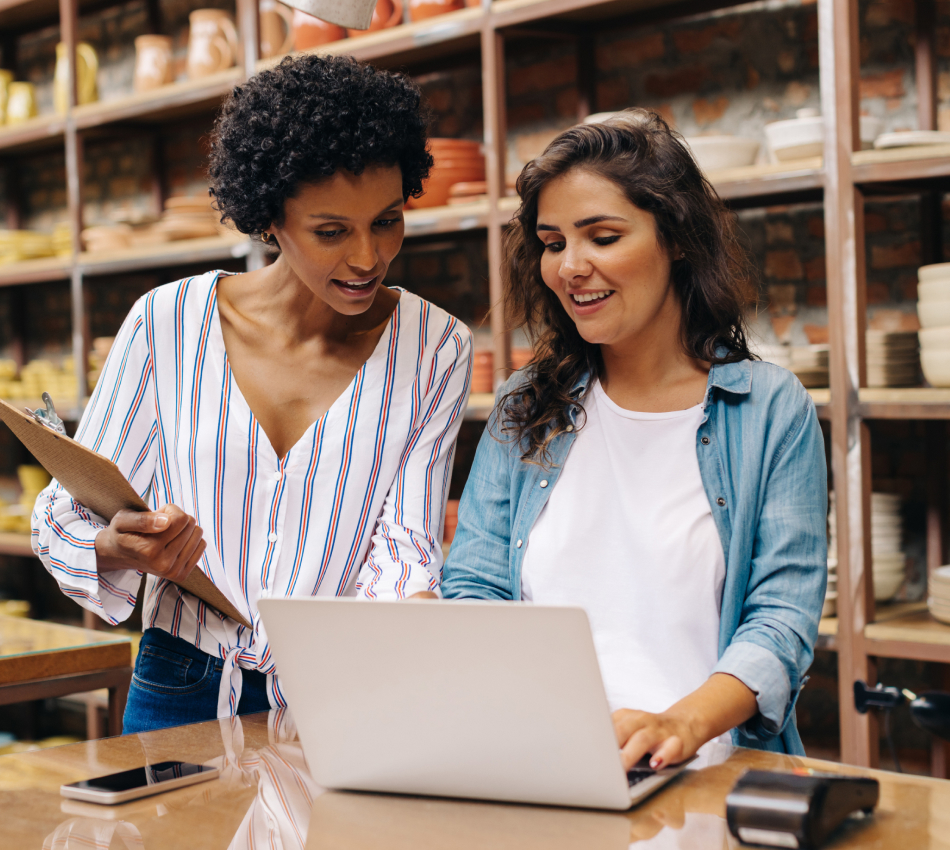 Two women strategize in a home goods store while one holds a clipboard and the other types on a laptop.