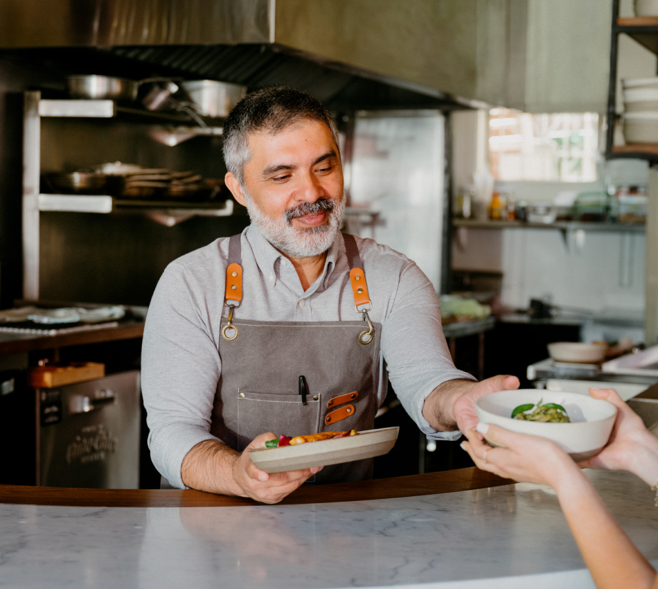 A male restaurant owner serves dishes to customers from behind the counter.